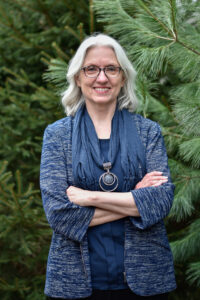 Dr. Cindy Fisher, arms folded standing in front of a pine tree. 